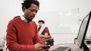 Como conseguir o primeiro emprego em TI - Foto de um homem preto usando o celular em frente ao computador